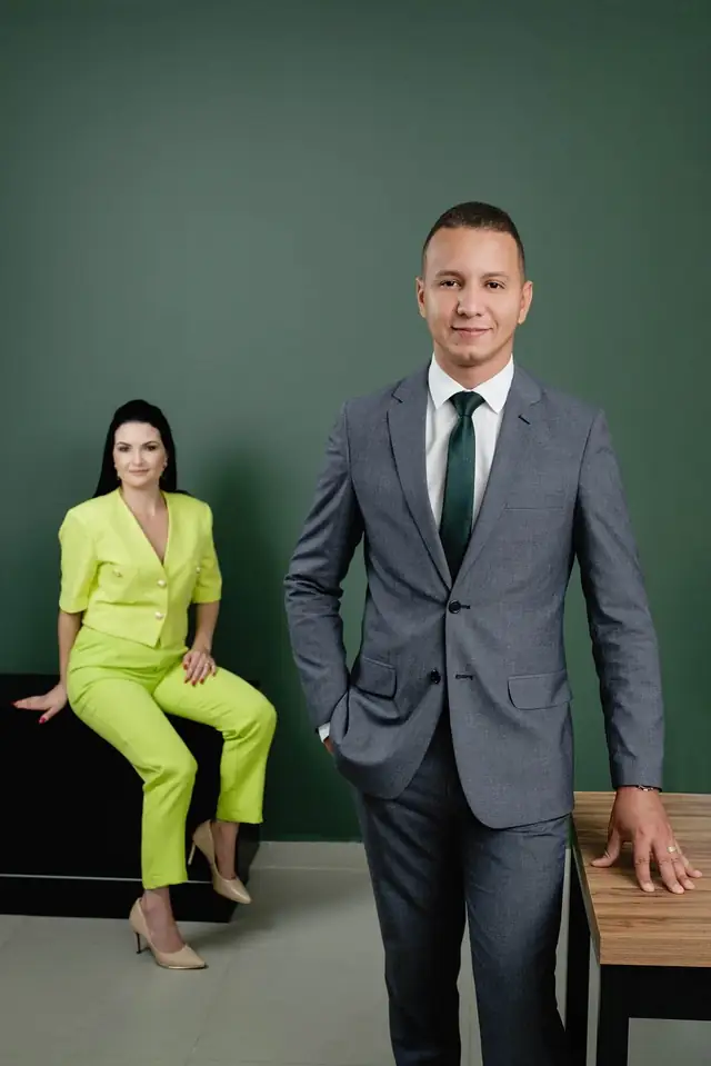 Os sócios no escritório: a advogada sentada, de conjunto verde-limão, e o advogado em pé, de terno cinza
